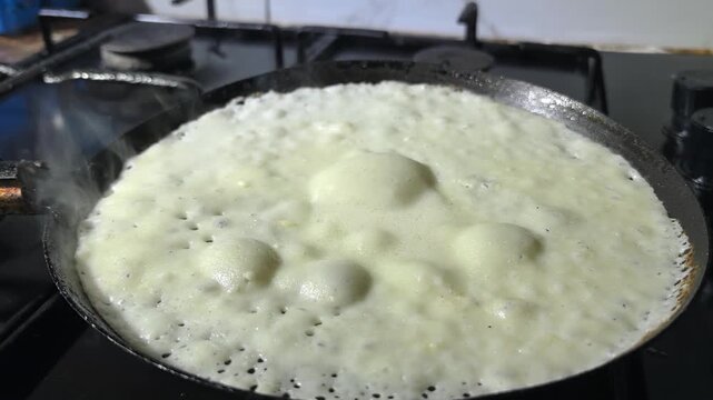 A dosa is being cooked on a hot pan. Bubbles are forming as the batter heats up. The kitchen shows a stove and cookware. This process involves careful timing.
