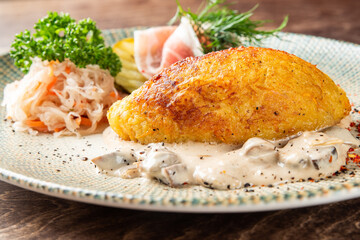 A hearty potato dumpling (likely cepelinai) served with creamy mushroom sauce, prosciutto, sauerkraut, and pickles on a rustic plate © pavel siamionov