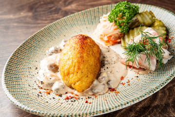 A hearty, fried potato dumpling (likely Cepelinai) is presented with creamy mushroom sauce, prosciutto, sauerkraut, and pickles on a patterned rustic plate © pavel siamionov