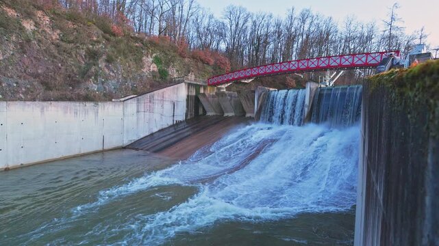Winter view of a dam with water flowing into a concrete chute. Red metal bridge on the left, calm lake on the right. Rocky cliff with forest in the background. Scenic landscape in Burgundy, France.