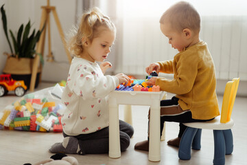 Naklejka premium Two young children sit together at a table playing with colorful building blocks. They focus on creating structures while surrounded by toys