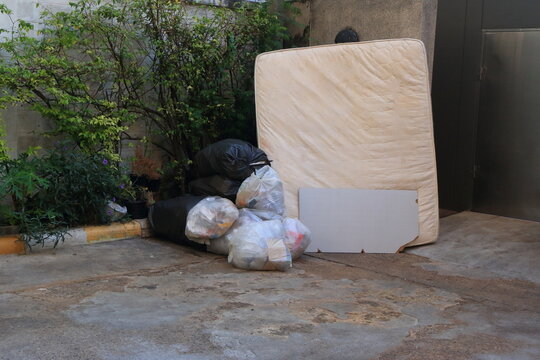 A discarded mattress rests against concrete wall near garbage room. The proper disposal of bulky items like this mattress involves specific municipal services or private companies to recycle it.