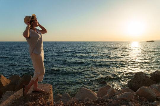 Full length of a young woman in a hat standing on the rocky shore, watching the sun set over the sea during summer vacation.