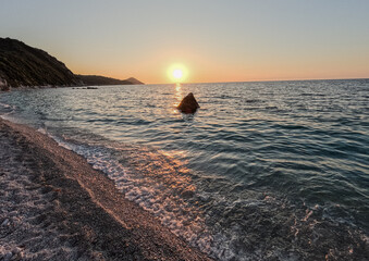 Sunset over a calm sea and rocky shore. A rock in the sea.