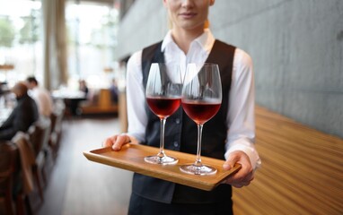 Professional waitress holding wooden tray with two glasses of rose or red wine, serving customers in elegant restaurant setting. Welcoming and elegant atmosphere for a special moment.