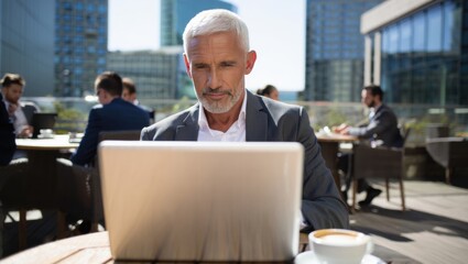 Focused mature entrepreneur using laptop and drinking coffee at an outdoor cafe in financial district. Senior businessman remote working, managing business online.
