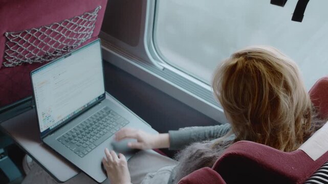 Woman working on a laptop while traveling by train, viewed from above. Remote work concept, modern business travel, focus, productivity, and mobility lifestyle.