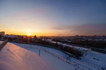 Panoramic winter cityscape viewed from a hill. A snow-covered highway interchange with sweeping curves leads toward a distant city of mid-rise buildings. To the right, a wide frozen river stretches al