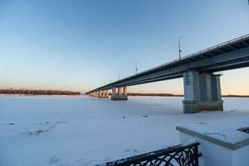 Fototapeta premium A massive steel road bridge stretches across a wide, frozen river under a clear blue winter sky. 