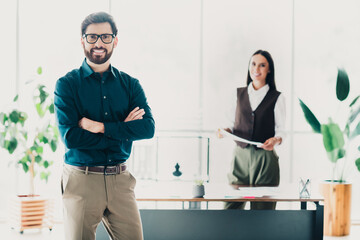 Obraz premium Confident businessman with arms crossed in bright modern office with friendly businesswoman in background