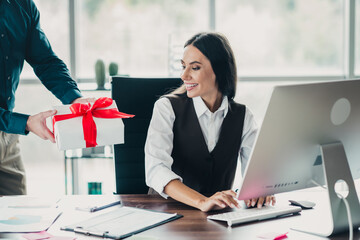 Businesswoman at a modern office desk receives a gift from a colleague while smiling at a computer...
