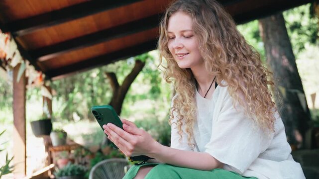 Young woman with long curly hair relaxing on a countryside terrace, scrolling social media on her smartphone during a warm summer day.