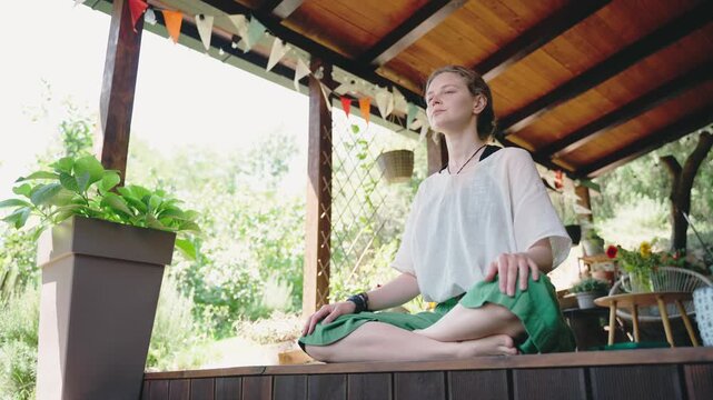 Young woman meditating on a countryside house terrace during a warm summer day. Wellness lifestyle, mindfulness, balance, relaxation, and peaceful outdoor living concept.