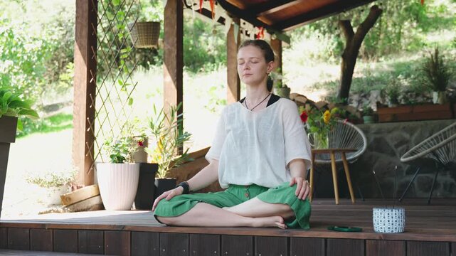Young woman meditating on a countryside house terrace during a warm summer day. Wellness lifestyle, mindfulness, balance, relaxation, and peaceful outdoor living concept.