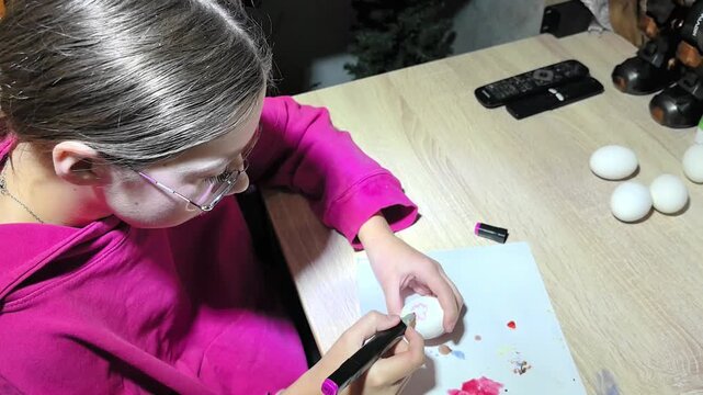 A child draws patterns on white eggs with a black marker. The scene takes place in a cozy room, where the child is decorating eggs for Easter.