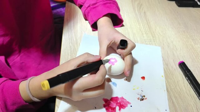 A child draws patterns on an egg lying on a table with a pink marker. Bright colors are scattered around the egg. The photo captures preparations for the Easter holiday.