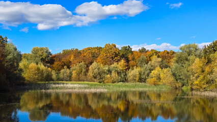 Beautiful autumn landscape in Romania with colorful trees reflecting on a tranquil lake under a blue sky with white clouds.