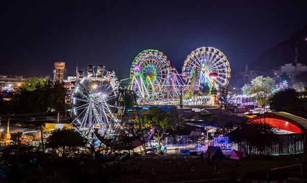 Colorful Ferris wheels and amusement rides glowing at night at a busy city fairground with festive lights.