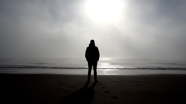 A solitary individual is silhouetted against a breathtakingly misty ocean horizon, where the sun attempts to break through the atmospheric haze, casting a soft, reflective glow on the calm water. Stan