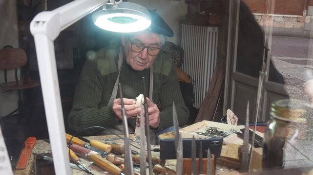 Skilled elderly craftsman meticulously carves a violin scroll in his detailed workshop