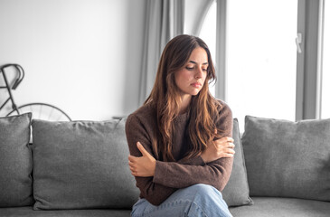 Obraz premium Portrait of serious and pensive young woman with long brown hair sitting on couch at home crossing arms
