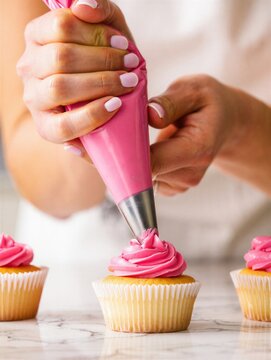 A baker is skillfully decorating freshly baked cupcakes with vibrant pink frosting using a piping bag. Delicious dessert preparation.