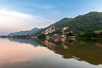 Obraz premium Blasting Mountains. View of mountain rock quarry by river under overcast sky. Landscape of riverside quarrying area with mountains and cloudy background. Environmental impact on mountain landscape.