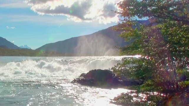 Wild waters erupt near the verdant shoreline. Mountains rise majestically in the background at Saltos del Petrohue. Sun illuminates the foamy rapids on a bright day in Chile.