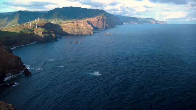 Drone low flight over the yellow rocky cliffs of Miradouro Ponta do Rosto, flying towards Machico region and the lush green mountains of Madeira eastern coast with wind turbines and Atlantic ocean.