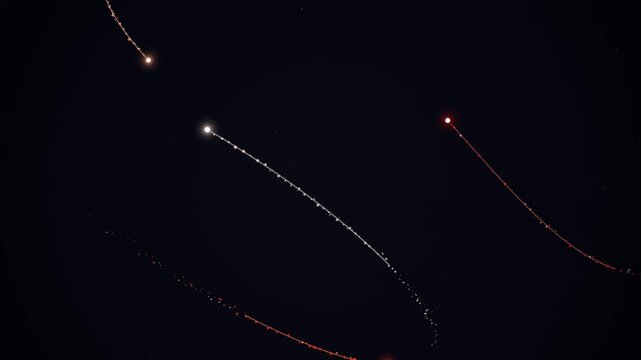 A nighttime scene with multiple glowing trails of light streaking across a dark sky with varying colors and intensities.