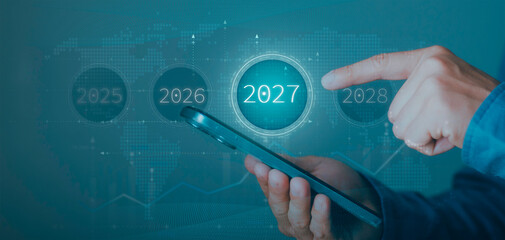 Businessman pointing at 2027 year icon on smartphone. Strategic future planning, business forecasting, long-term goals, and financial roadmap on global world map and graph background. © Krittamet