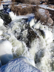 Winter waterscape. Frozen waterfall, shore with reeds, sunny day landscape, bubbling water stream, ice covered granite rocks, snow carpet, icicles. Freathing cold winter weather in February. 