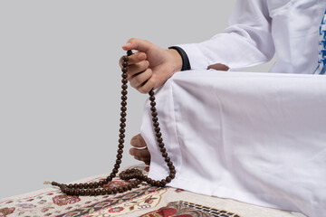 Arab man kneeling on prayer rug, wearing white thobe and kufi, holding wooden prayer beads, closeup of hands and beads, neutral