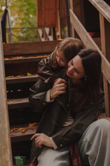 Mom and daughter are sitting on a wooden staircase. Mom and daughter are playing. A house with a green roof on the background. Mothers Day. The girl climbs to hug her mother. Vertical photo
