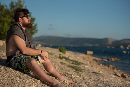 Side view of a shirtless young man with a towel over his shoulders sitting on a pebble beach looking out to sea, Brodarica, Sibenik, Dalmatia, Croatia