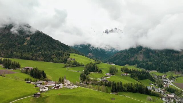 4K aerial of St. Magdalena (Santa Maddalena), Val di Funes, Dolomites, South Tyrol, Italy&mdash;alpine church, green meadows and farms under moving fog and low clouds. Captured May 5, 2025.