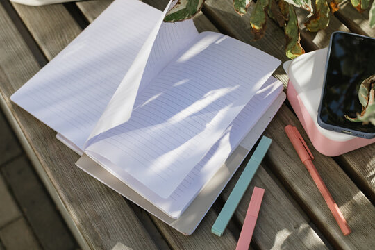 Overhead view of an open blank notebook, pens, a mobile phone and lunch box on a park bench in the sunlight