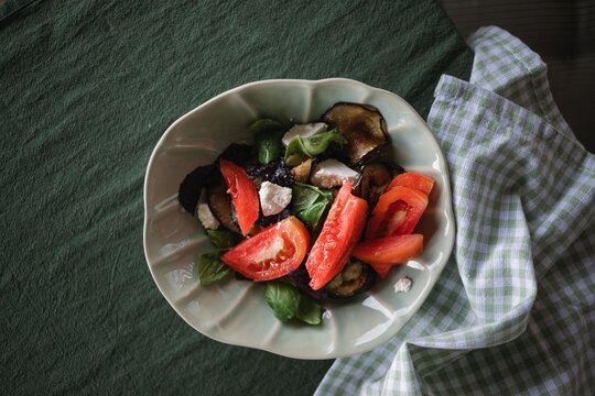 Overhead view of a bowl of fresh tomatoes, grilled eggplant, feta cheese, and basil leaves drizzled with olive oil on a table with a napkin