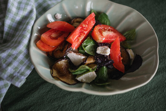 Overhead view of a bowl of fresh tomatoes, grilled eggplant, feta cheese, and basil leaves drizzled with olive oil on a table with a napkin