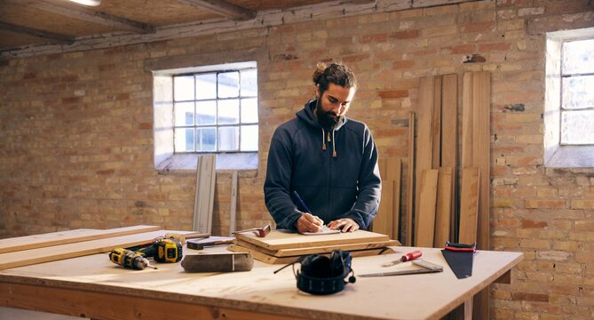Woodworker drawing designs at a bench in his studio. Tools are placed beside him.