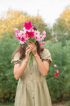 Close-up of a young Woman standing in a  garden holding a bunch of fresh pink cosmos flowers in front of her face