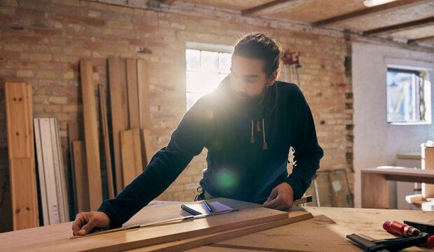 Woodworker measuring a plank on a bench. Golden rays of sunlight pour in through the window behind him. Tools are placed beside him.