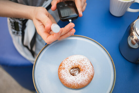 Close-up overhead view of a young woman with type 1 diabetes sitting at a cafe table with a doughnut and coffee checking her insulin levels with a glaucometer