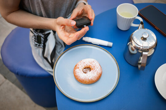 Close-up of a young woman with type 1 diabetes sitting at a cafe table with a doughnut and coffee checking her insulin levels with a glaucometer