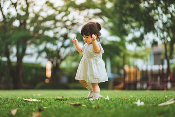 Cute Asian toddler girl in white dress walking on green grass in a sunny park. Childhood first steps exploration and outdoor play concept. Little girl enjoying nature on weekend