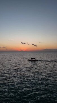 Aerial drone view of a lone fisherman rowing his small wooden boat during sunset in Vlora Bay, Albania, with soft orange skies, calm waters, and distant silhouettes of hills and clouds