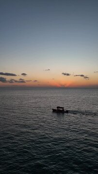 Aerial drone view of a lone fisherman rowing his small wooden boat during sunset in Vlora Bay, Albania, with soft orange skies, calm waters, and distant silhouettes of hills and clouds
