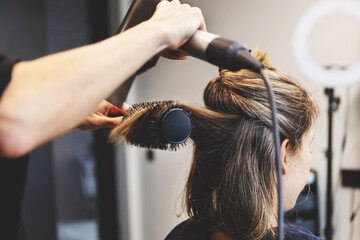 Fototapeta premium Hairdresser blow drying hair of woman with round brush in modern beauty salon, styling sectioned strands. Close up professional hairstyling