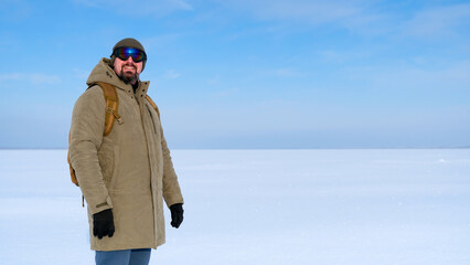 Man with beard in beanie, ski goggles and winter jacket smiles at camera while standing on a vast snow covered field under a clear blue sky, evoking outdoor adventure