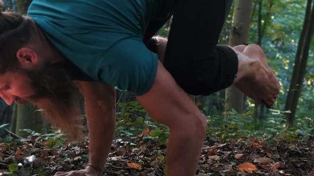 Close-up of muscular man doing crow pose (bakasana) and pushups on forest floor, strength and balance outdoor yoga practice.